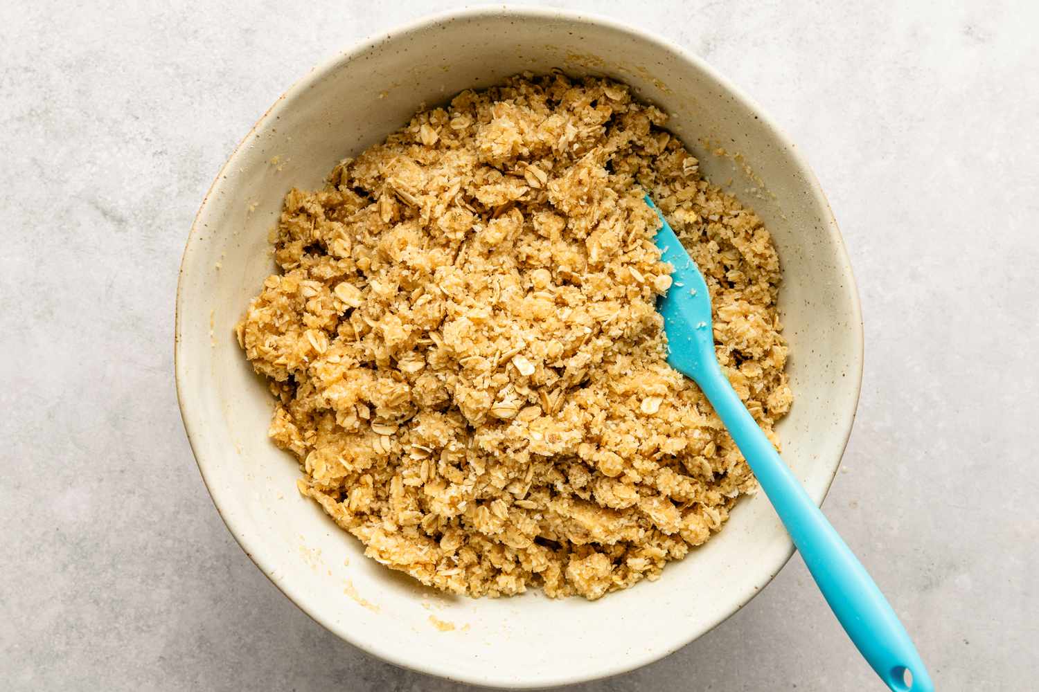 Dry and wet ingredients for the Anzac Biscuits recipe getting mixed together with a spatula in a white bowl