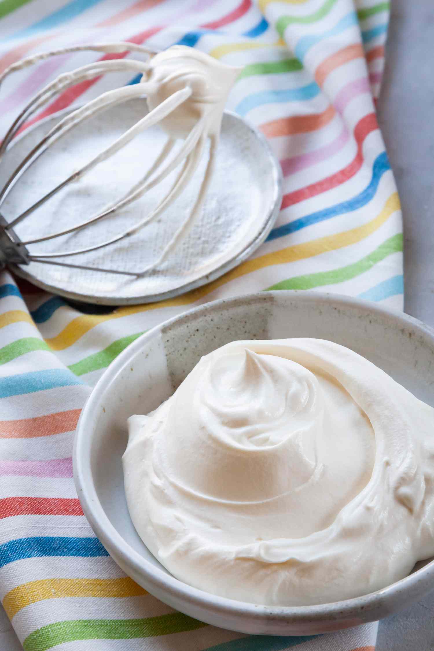 Whipped cream frosting with cream cheese in a bowl with a whisk next to it and a striped linen underneath.