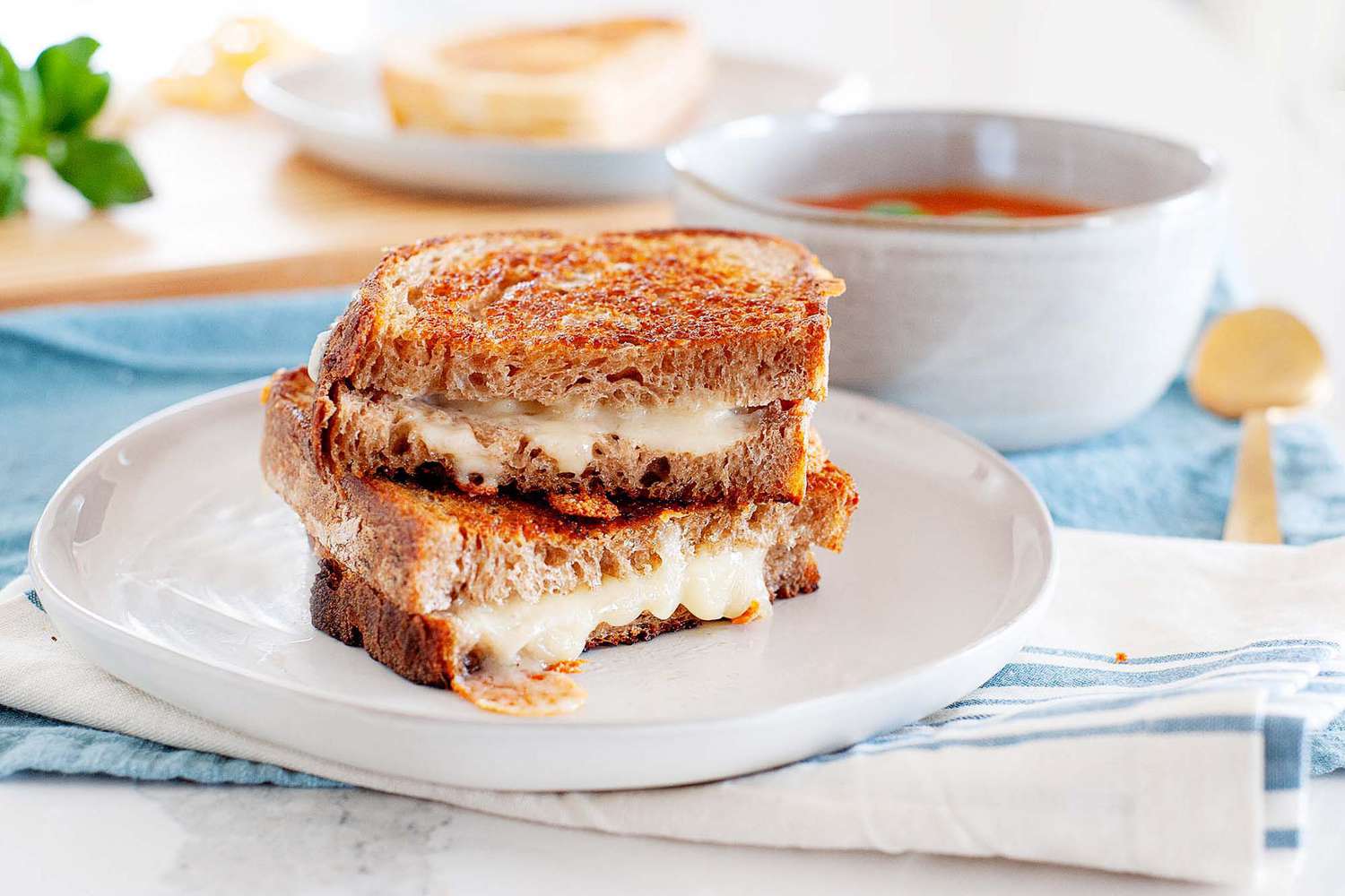 The perfect grilled cheese sandwich cut in half and split open on a plate with a blue napkin underneath it and a bowl of tomato soup in the background.