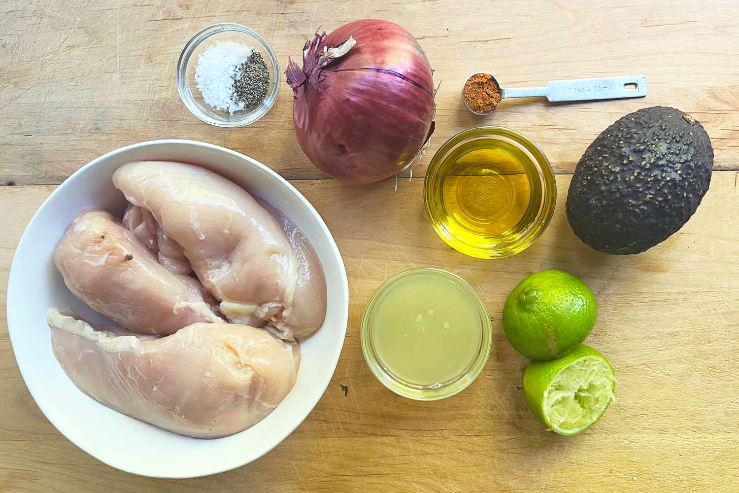 Ingredients including raw chicken red onion avocado lime oil cayenne and salt and pepper arranged on a wooden surface