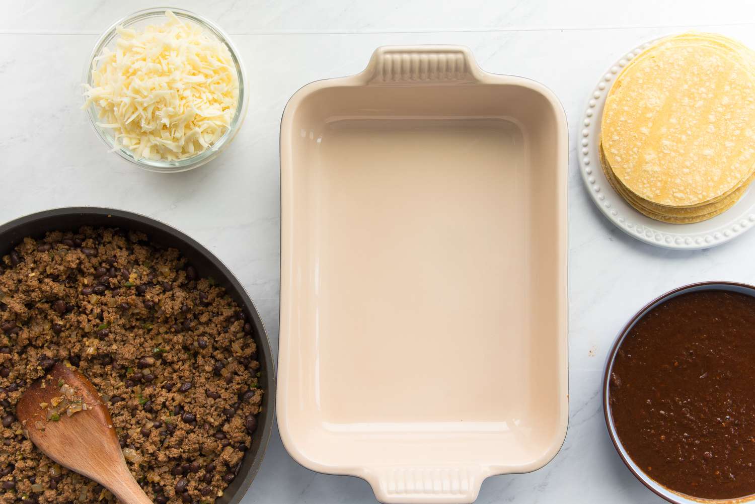 Casserole Dish Surrounded by Beef Enchilada Casserole Ingredients (Pan of Cooked Beef, Bowl of Shredded Cheese, Plate with Corn Tortillas, and a Bowl of Red Enchilada Sauce)
