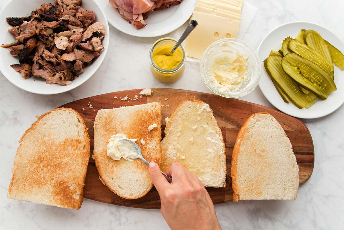 A wooden cutting board is set on a marble background. French bread is cut into quarters and toasted. A hand is using a spoon to spread mayonnaise on the center piece of bread. Bowls of shredded pork, sliced ham, yellow mustard, sliced swiss cheese, mayonnaise and sliced pickle are set above.