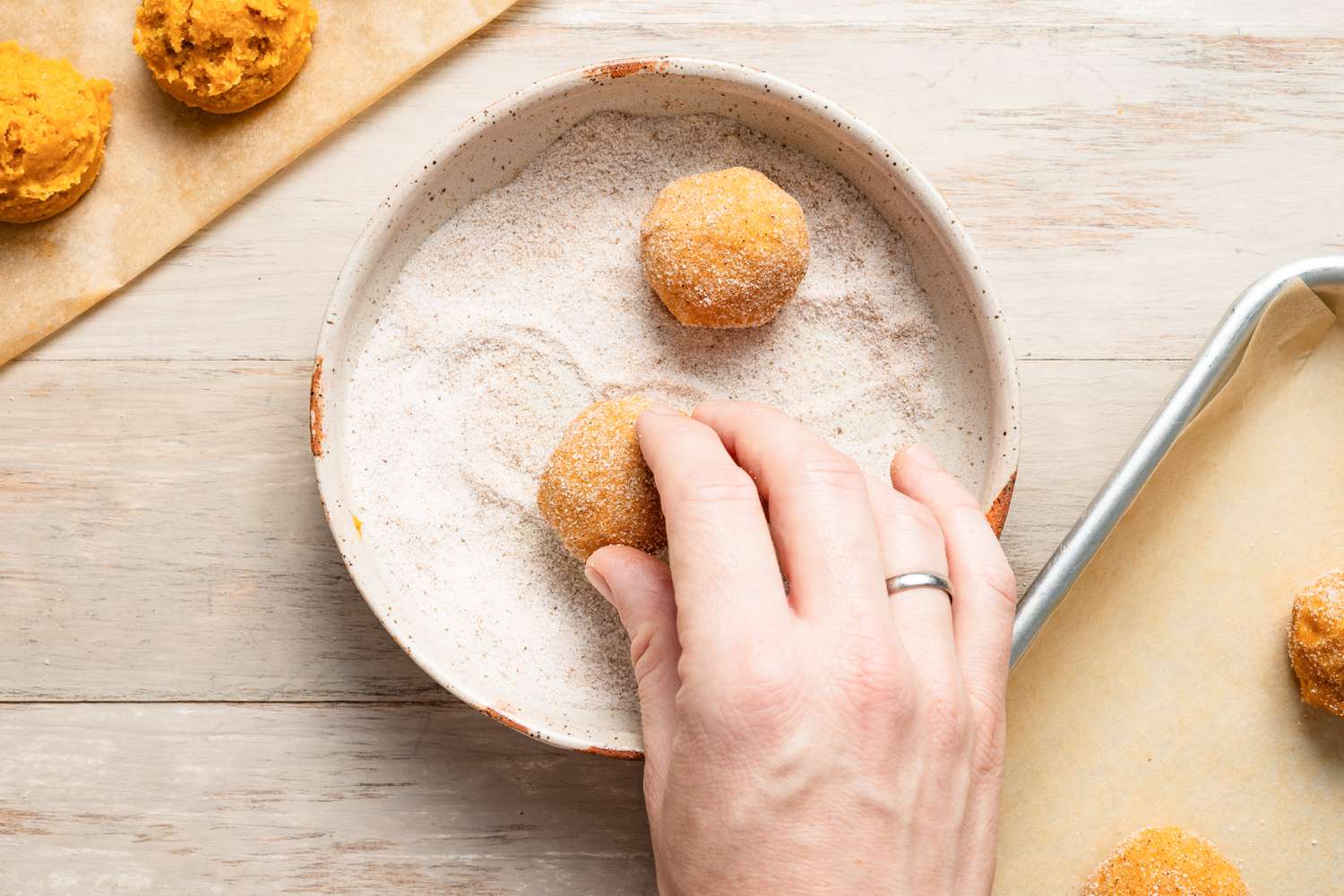Hand rolling balls of snickerdoodle pumpkin spice cookie dough rolled in a bowl of pumpkin spice sugar mixture. To the left of the bowl, more pumpkin spice cookie dough balls, and to the right of the bowl, a parchment paper lined baking sheet with sugar covered cookie dough balls.