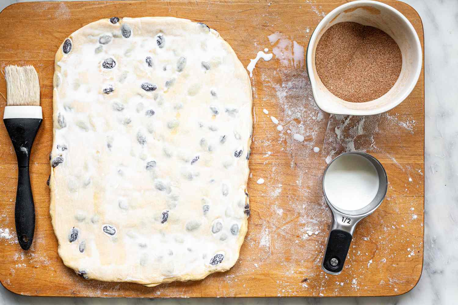 Cinnamon Swirl Dough Patted Flat into a Rectangle on Wooden Cutting Board and Brushed with Milk. Next to the Dough, a Bowl of Cinnamon Sugar and a 1/2 Measuring Cup with Milk 