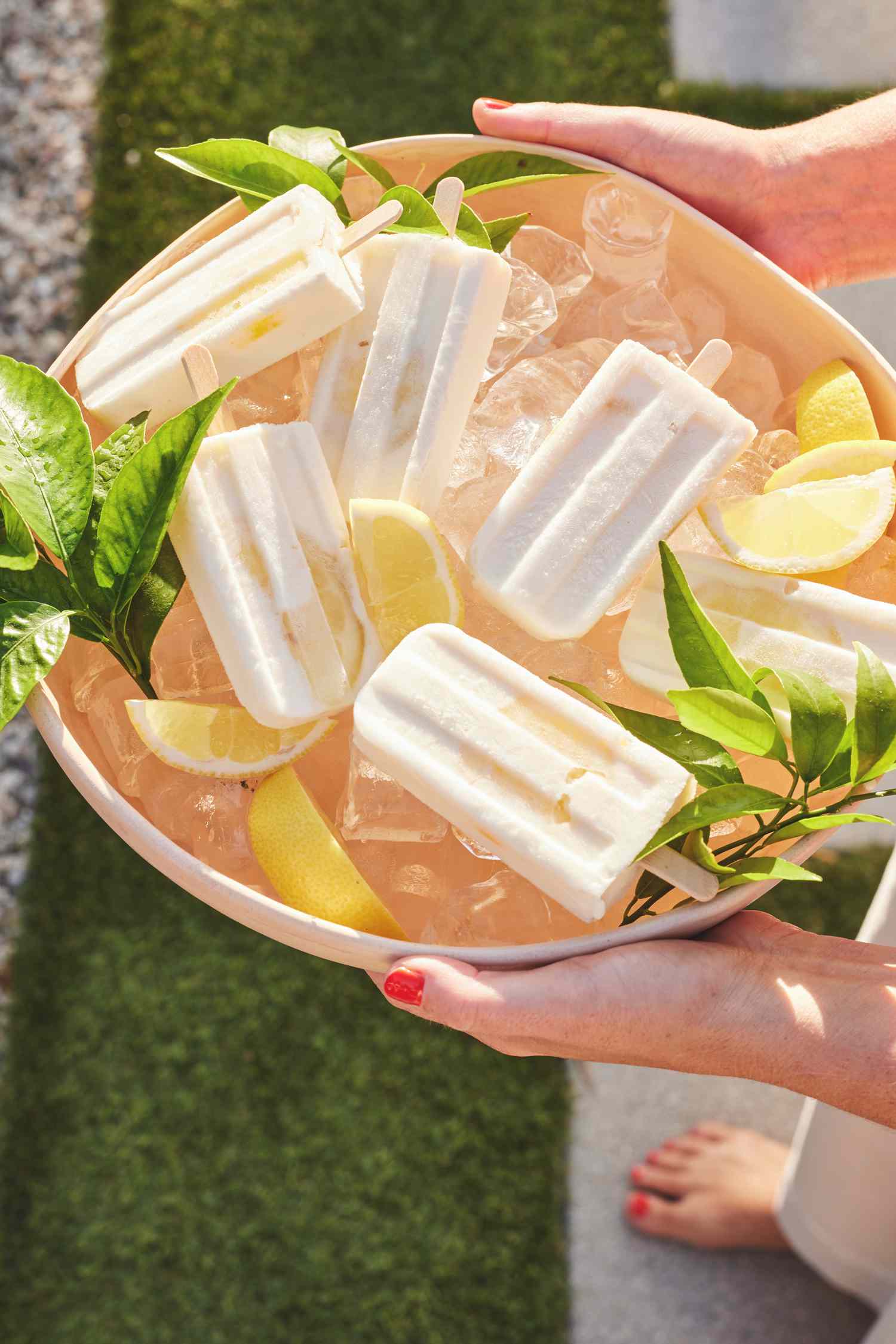 Someone Holding a Bowl of Limoncello Yogurt Pops on Ice Surrounded by Leaves and Lemons Over Grass