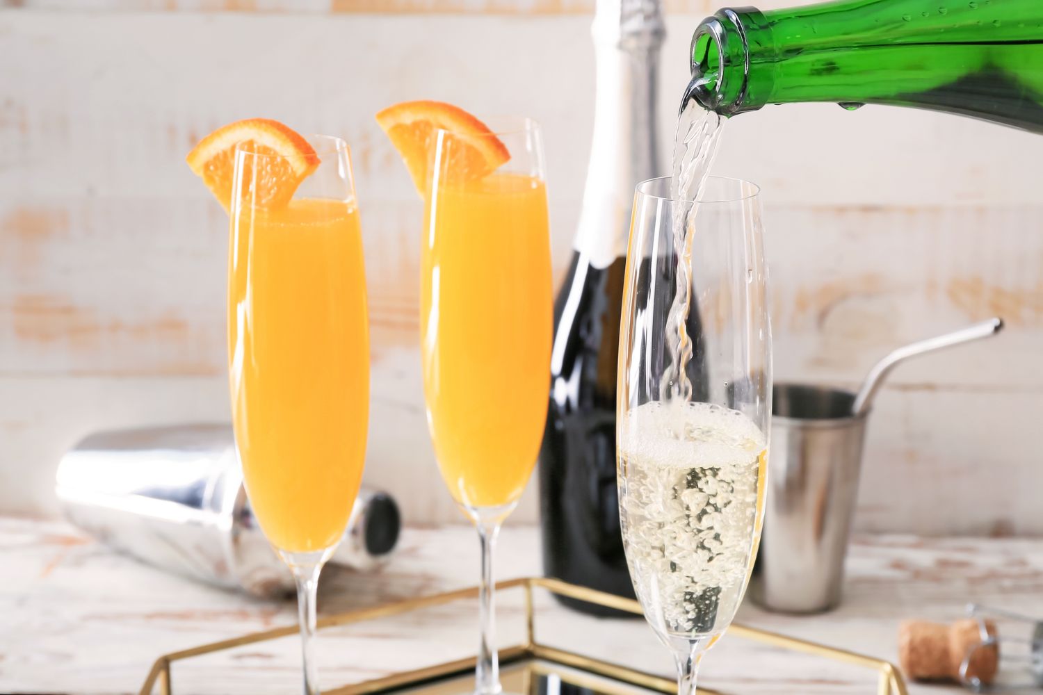 Side view of a two tall glasses of Mimosa cocktails with an orange slice on the rim next to a tall glass being filled with sparkling wine from a green wine bottle