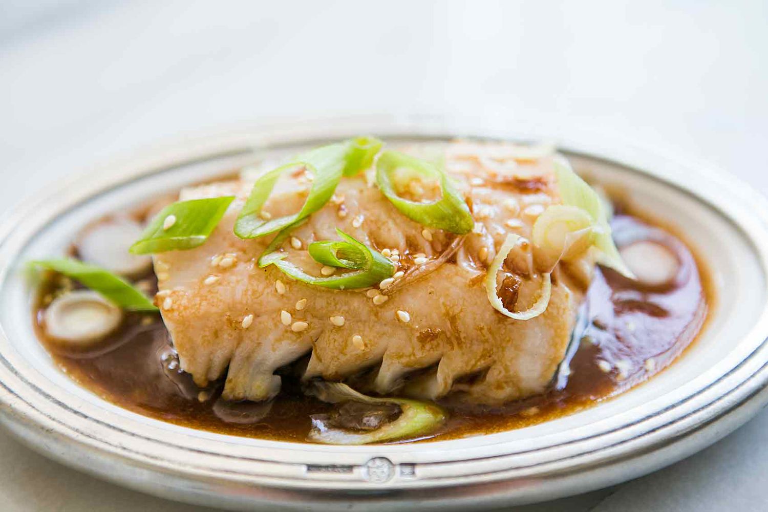 A dish featuring a piece of cooked cod garnished with green onions and sesame seeds served in a brown sauce on a plate
