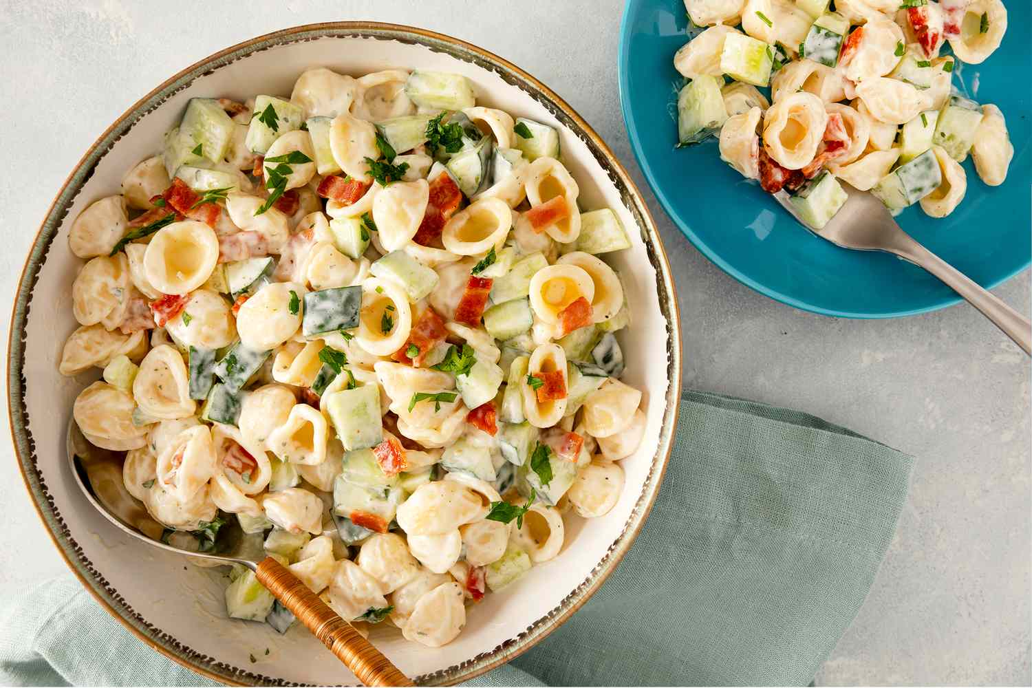 A bowl and a plate of pasta salad with cucumbers bacon and a ranch dressing on a table with a napkin