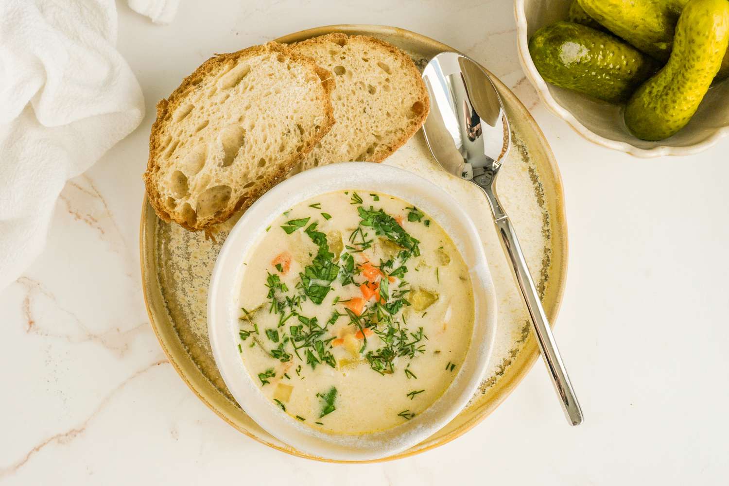 A bowl of soup garnished with herbs accompanied by slices of bread and a side of pickles
