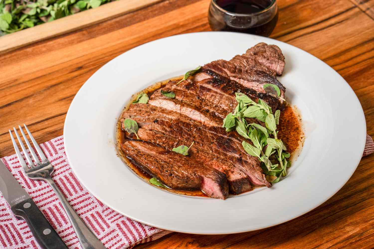 Sliced steak served on a white plate with garnishing, next to dining utensils and a glass of wine on a wooden table