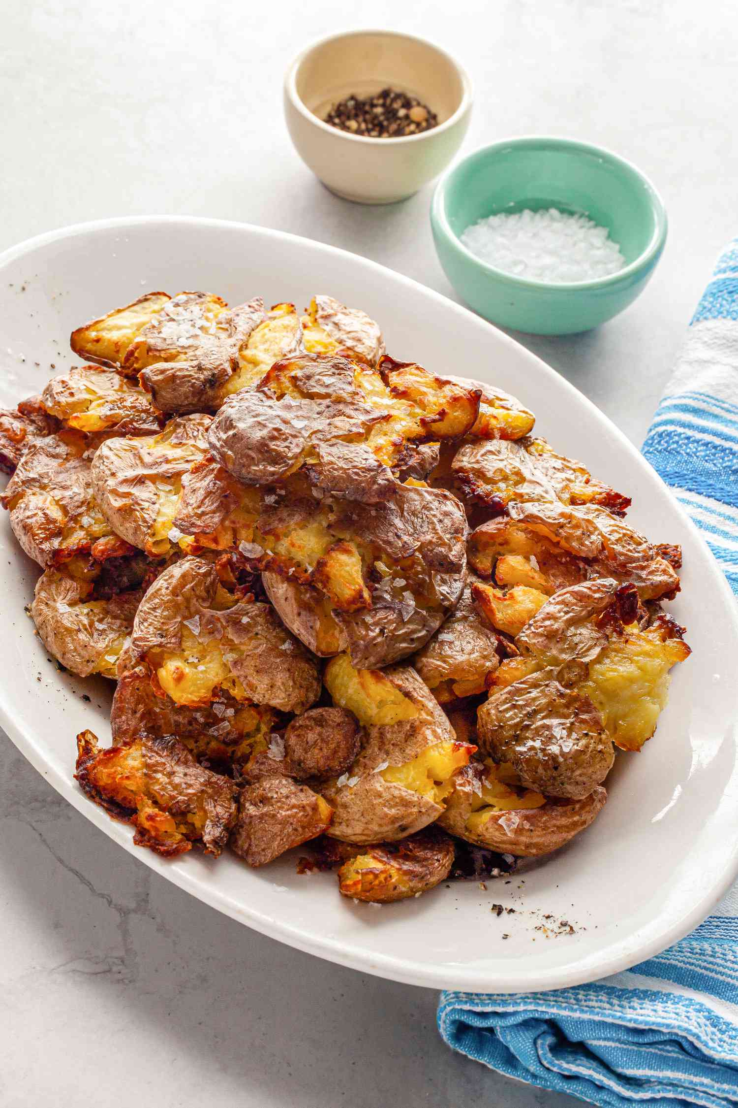 Smashed Potatoes on a Plate, and Next to It, Two Small Bowls (One with Cracked Pepper and Another with Flaky Salt)
