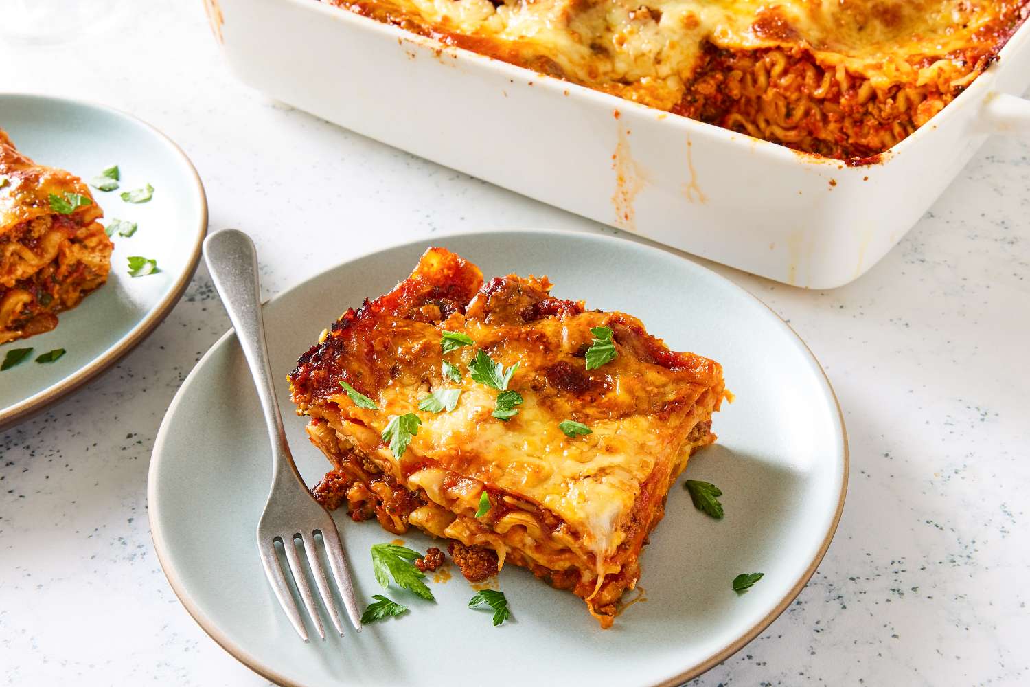 A serving of homemade lasagna on a plate with a fork casserole dish of lasagna in the background