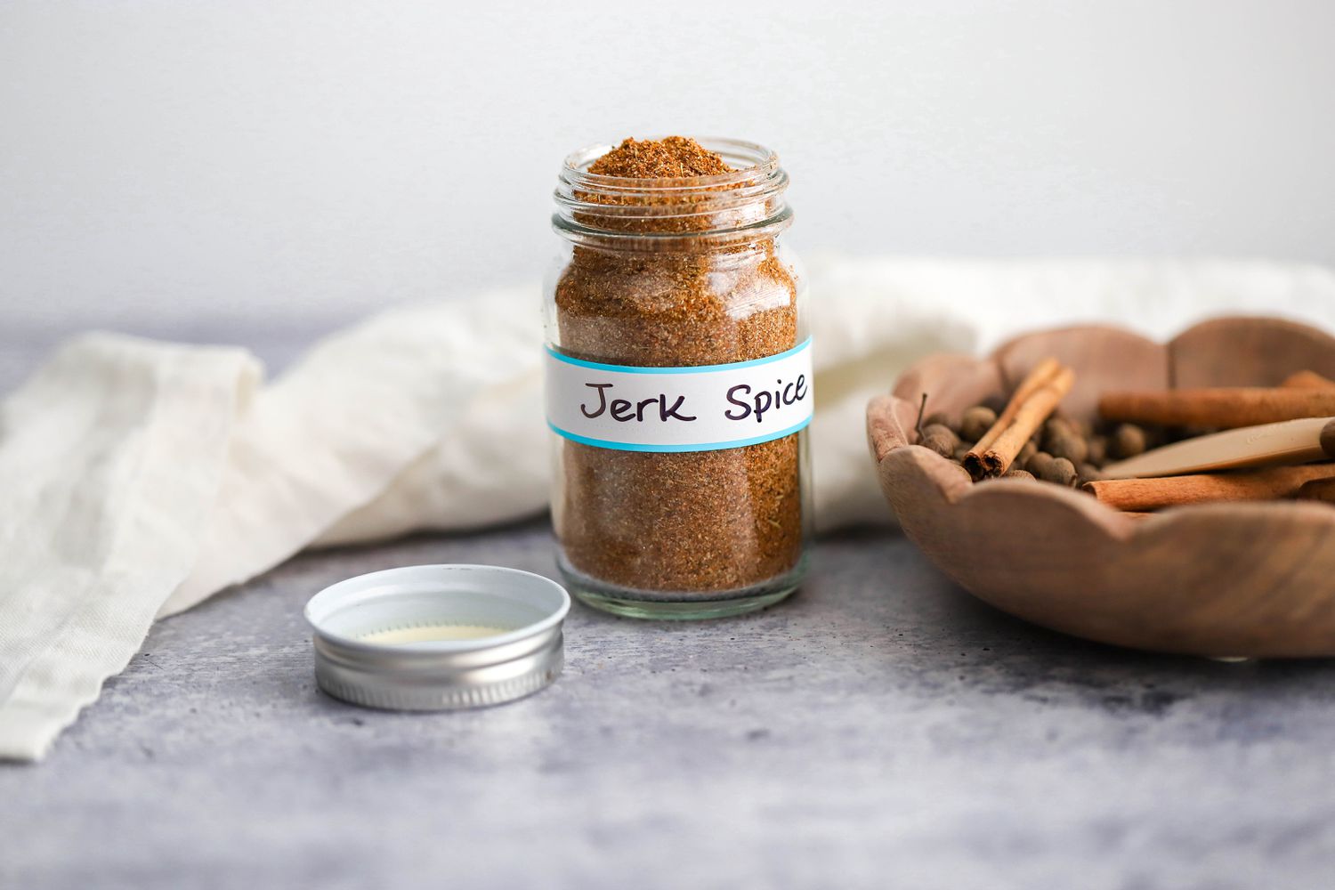 Jar of Jerk Seasoning With a Label Reading "Jerk Spice", and in the Surroundings, a Kitchen Linen and a Bowl With Whole Allspice and Cinnamon Sticks