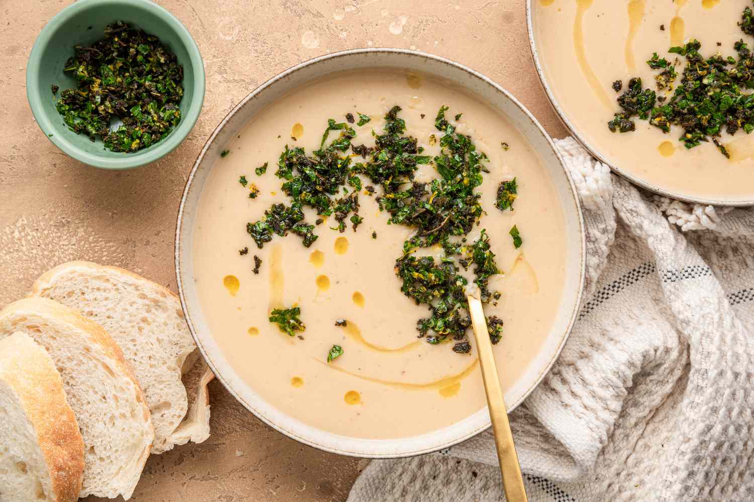 Bowl of Creamy White Bean Soup Topped with Sage Gremolata, Surrounded by Slices of Bread on the Counter, a Bowl of Sage Gremolata, and Another Bowl of Soup