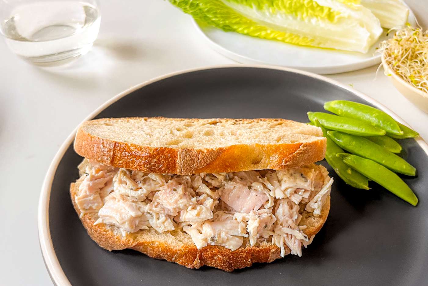 2-ingredient chicken salad sandwich on a plate with snap peas, and in the background, a plate with lettuce and a bow of microgreens
