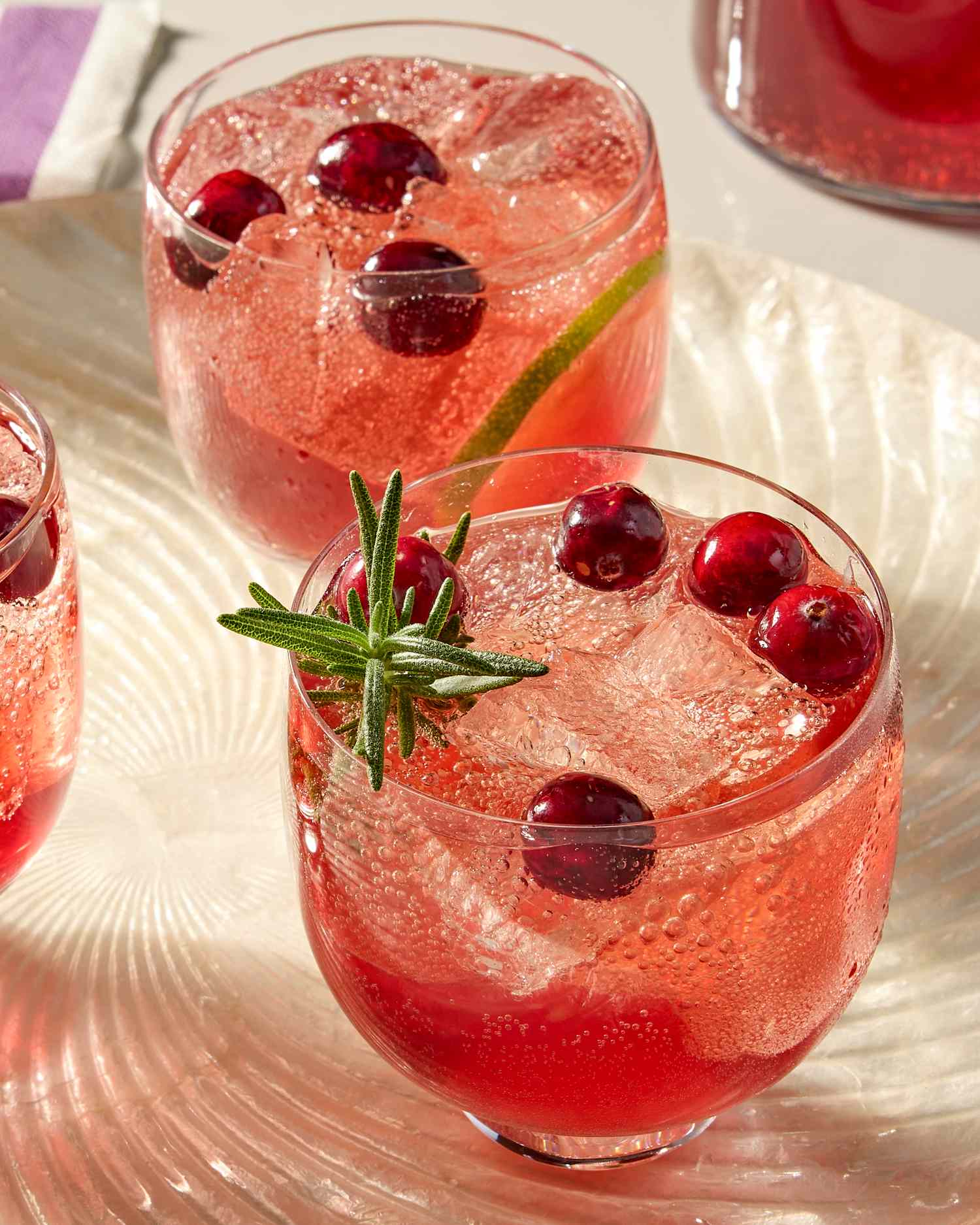 Two glasses of cranberry ginger spritz garnished with cranberries lime slices and rosemary served on a textured surface