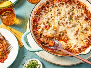 unstuffed peppers in a skillet at a table setting with a serving on a plate, a glass of water, a small dish with minced herbs, and a stack of plates