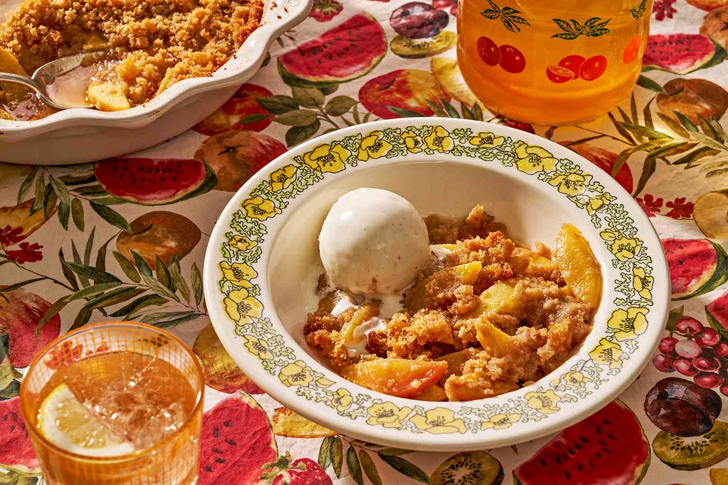 bowl of peach brown betty with a scoop of ice cream next to a pie dish with more and ice tea in two glasses, all at a cozy table setting (tablecloth pattern is watercolor cut fruit)