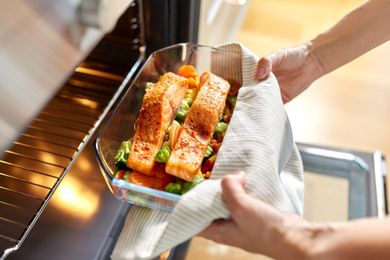 Hands placing a glass dish with roasted salmon and vegetables into an oven