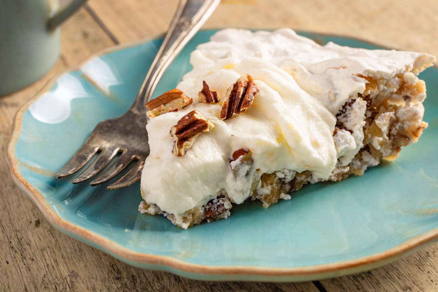 Closeup view of a slice of cream pie topped with pecans on a turquoise colored plate along with a fork all on a wooden tabletop