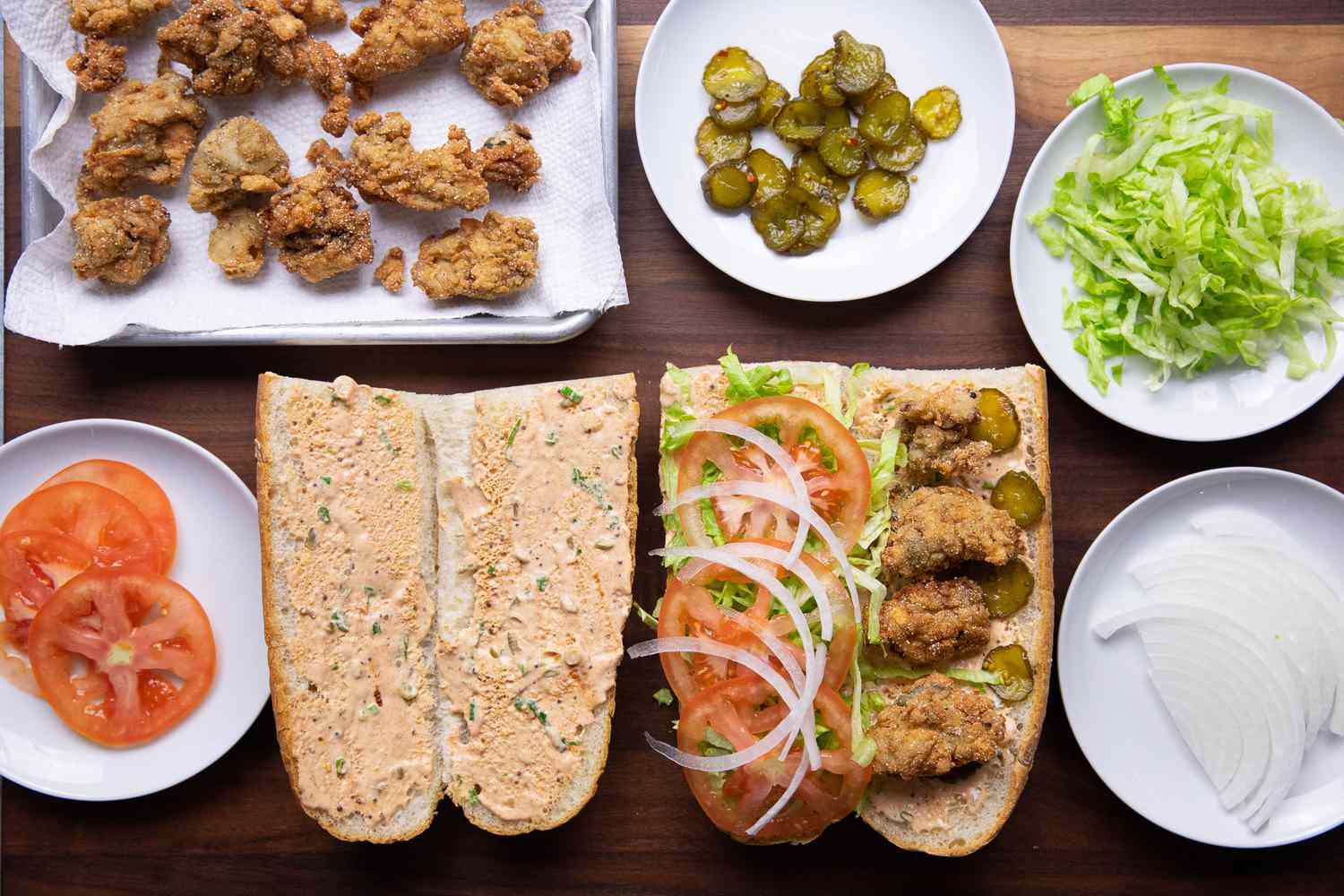 Assembling the Oyster Po' Boy (from L to R): Fried Oysters on a Paper Towel Lined Baking Sheet, Small Plate with Sliced Tomatoes, Small Plate of Pickles, French Bread Smeared with Remoulade, Another Loaf with Oyster Po Boy Ingredients, Small Plate with Shredded Lettuce, and Small Plate with Sliced Onions