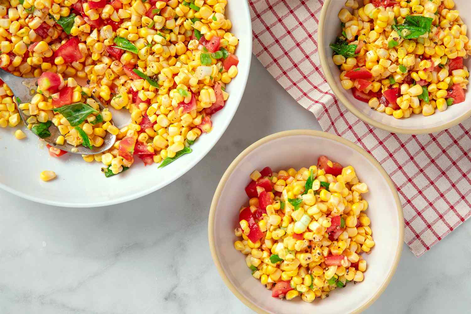 Bowls and a plate of corn salad with tomatoes and basil on a table with a checkered cloth