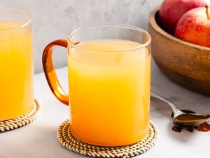 Two mugs of apple cider on coasters a wooden bowl with apples and a spoon with caramel nearby