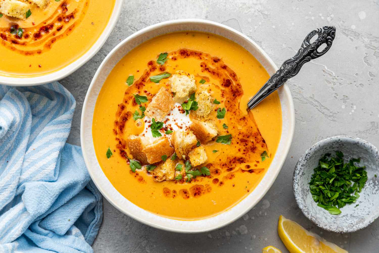 Bowls of Mercimek Corbasi (Turkish Lentil Soup) Topped with Greek Yogurt, Croutons, Pul Bibir Butter, and Parsley, and Next to It, a Small Bowl of Parsley and Lemon Wedges