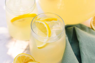 Two Glasses of Lemonade With Ice and Lemon Slices Next to a Pitcher With More Lemonade, Halved Lemons on the Counter, and a Sage Table Napkin 