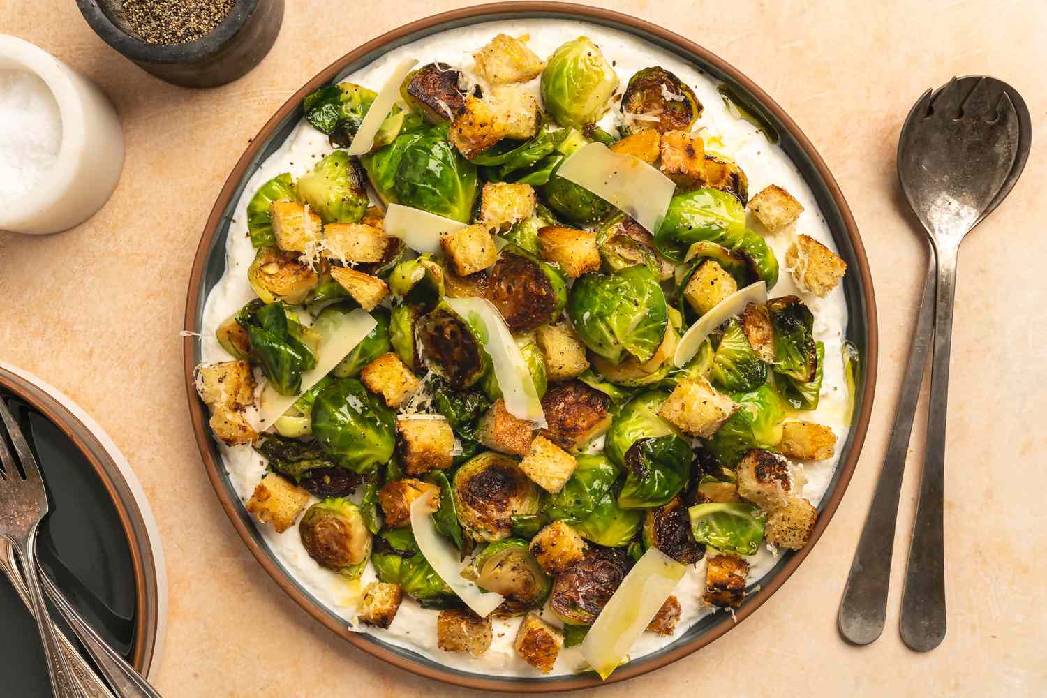 A plate of roasted Brussels sprouts with croutons and shaved cheese presented on a dish with a creamy base utensils nearby