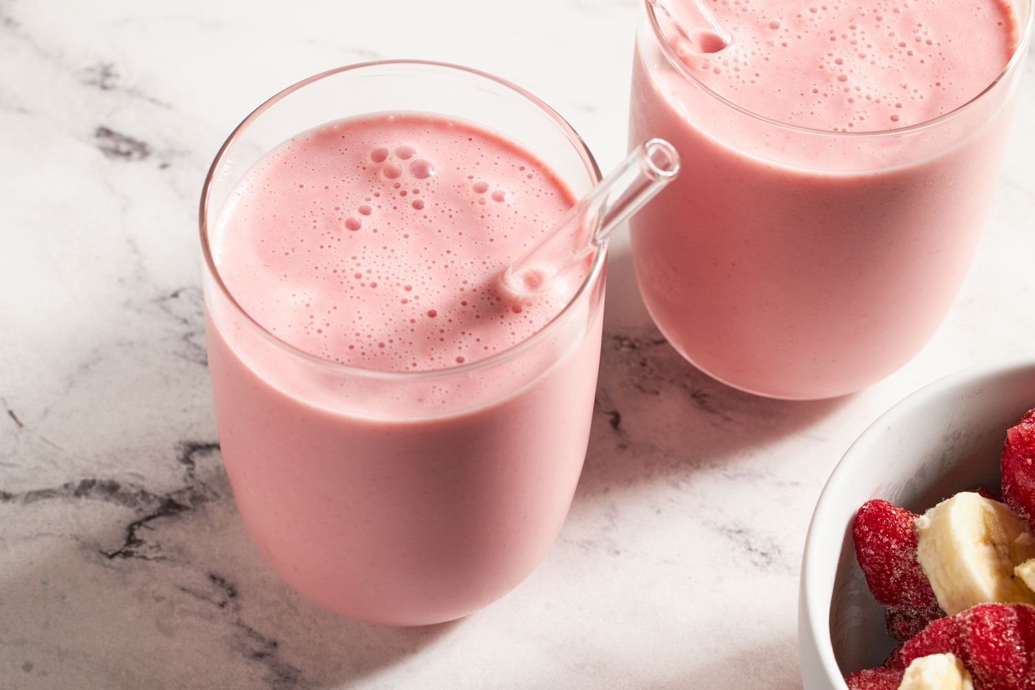 Two glasses of pink smoothie with glass straws next to a bowl of sliced bananas and frozen raspberries