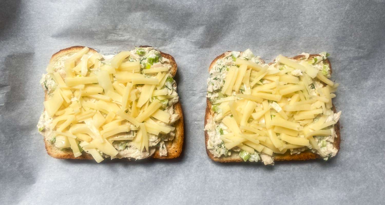 Two slices of bread topped with shredded cheese and tuna mixture, arranged on a baking sheet