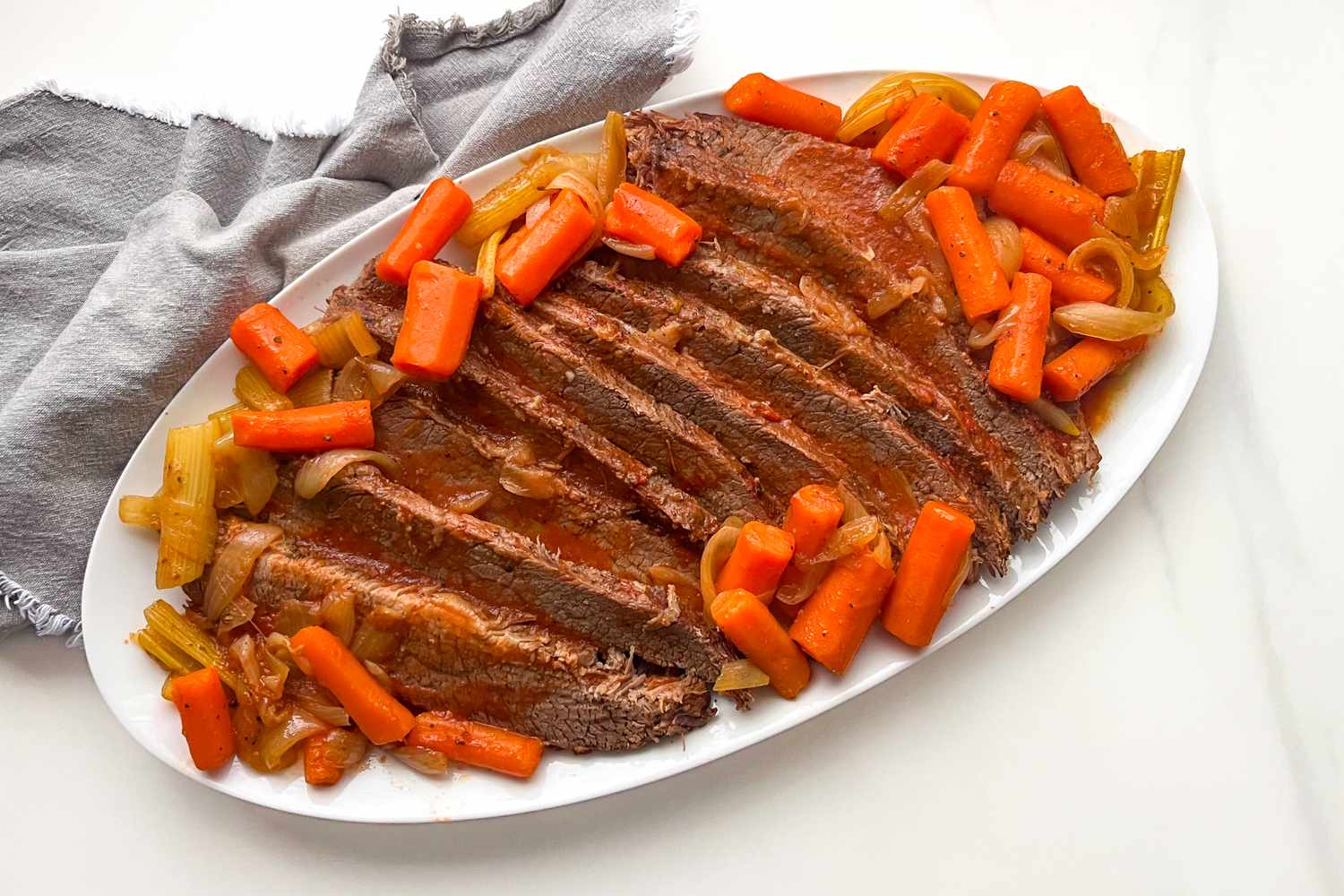 Sliced brisket served on a white platter with cooked carrots onions and celery