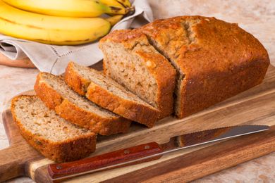 A partly sliced banana bread on a cutting board, with bananas in the background