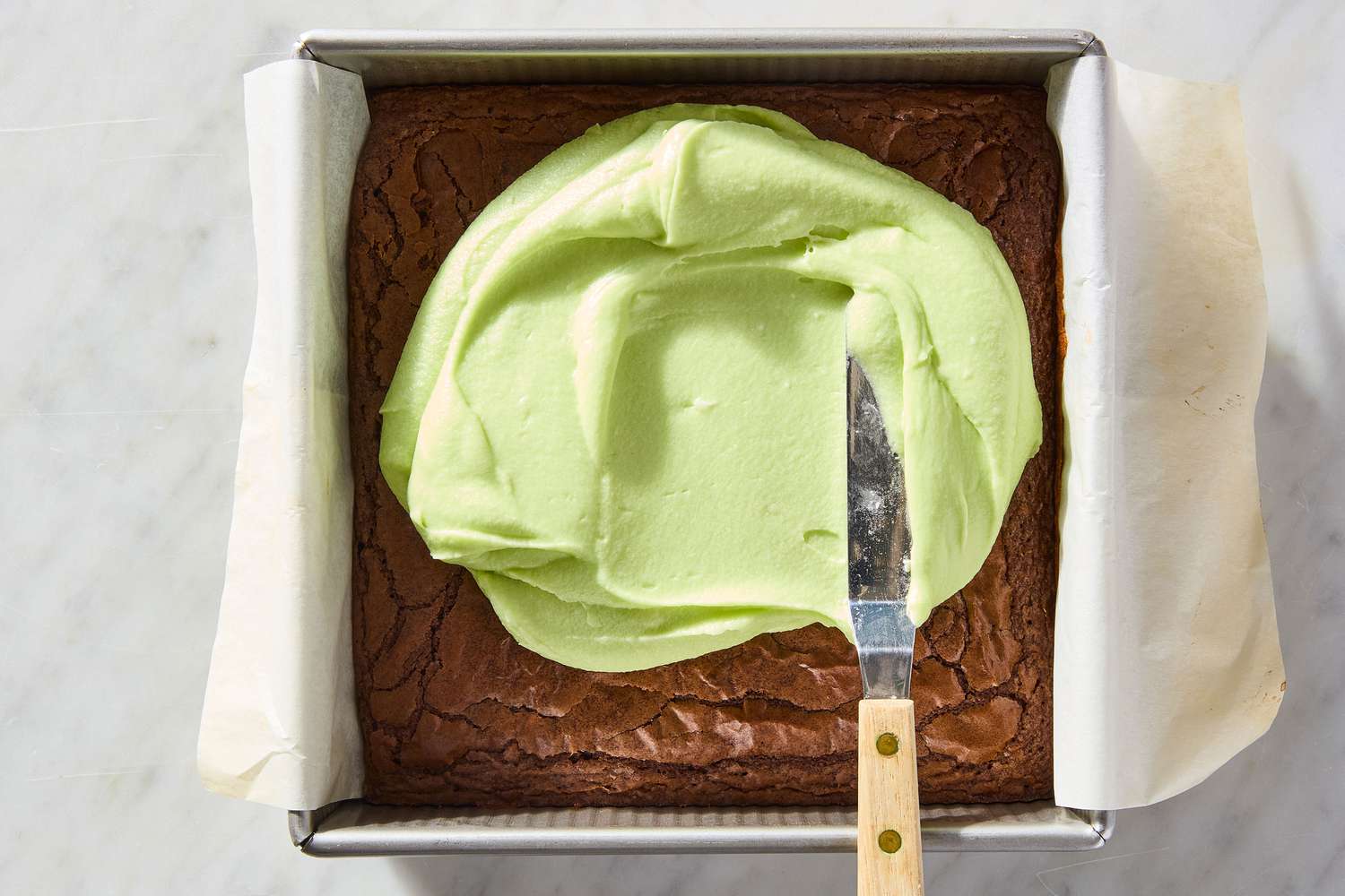 overhead view of knife spreading mint mixture on top of cooked brownie in pan for Mint Brownies