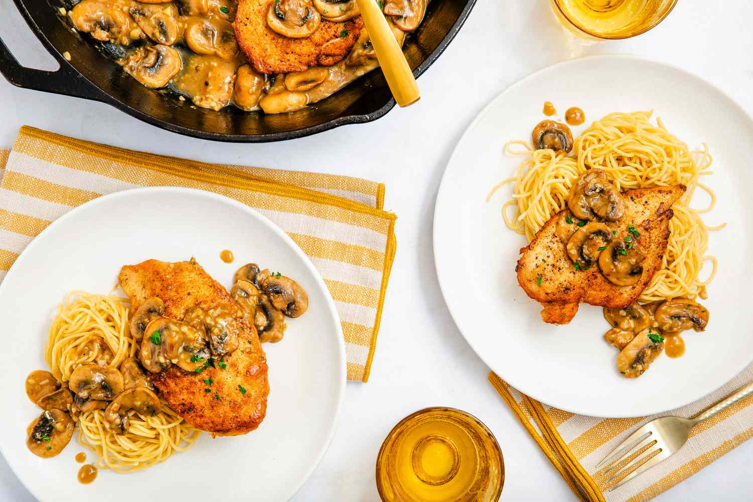 chicken marsala served with spaghetti (two plates) at a table setting with a table napkins, glasses, and more chicken marsala in the cast iron skillet