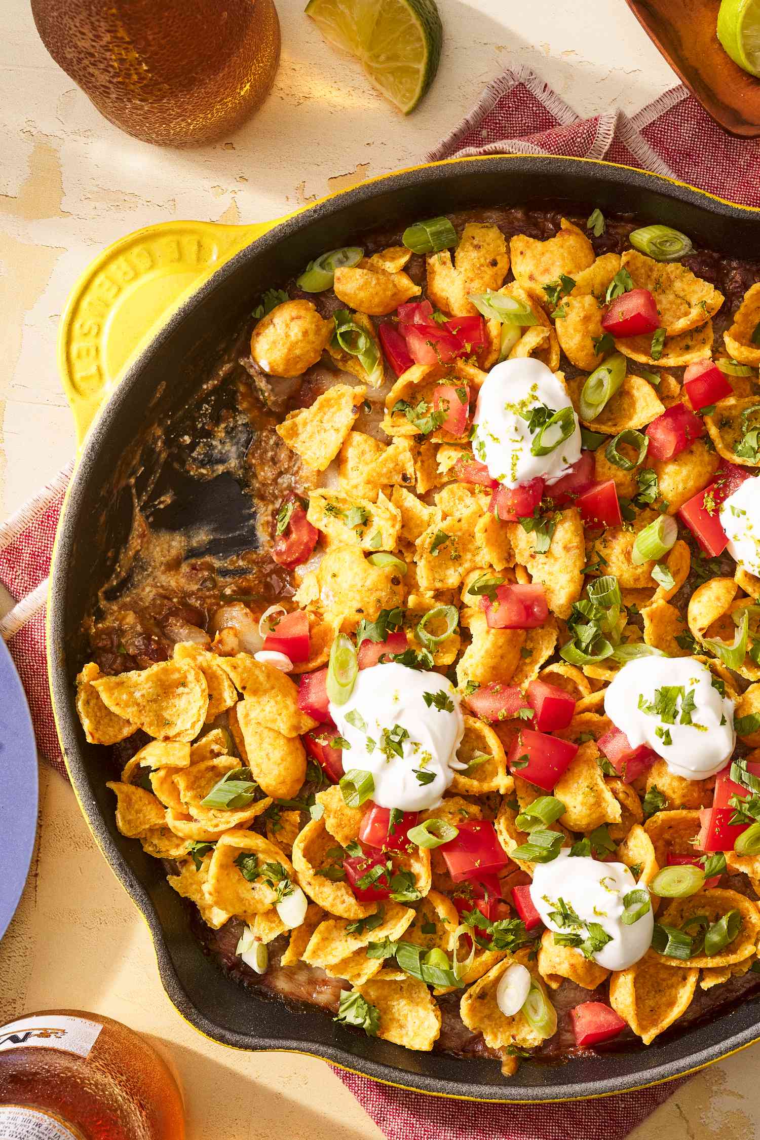 Frito pie dip in a cast iron skillet at a table setting with beer, plates, and lime wedges