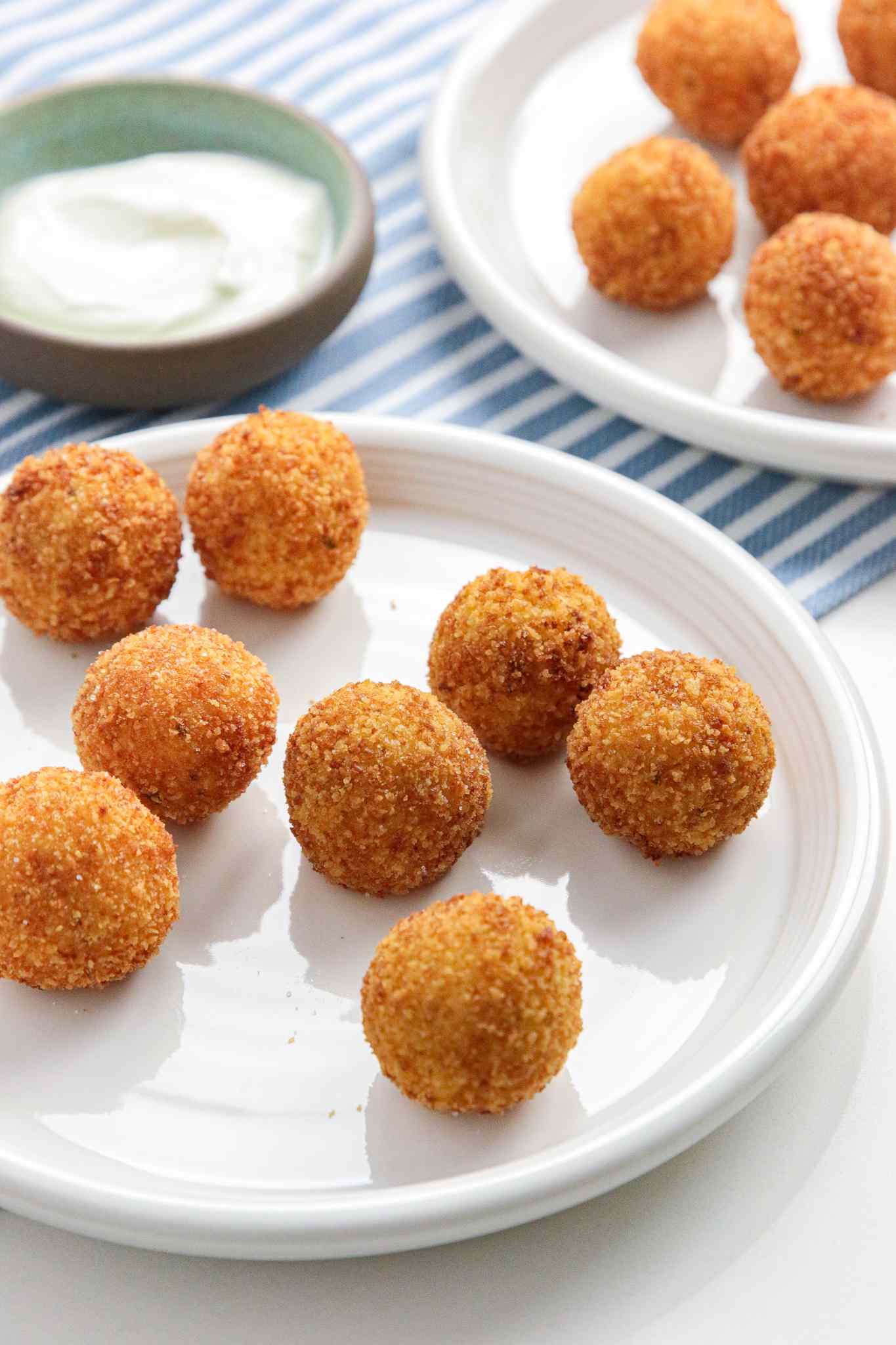 Two Plates of Potato Croquettes Next to a Small Saucer of Sauce on a Table Cloth 