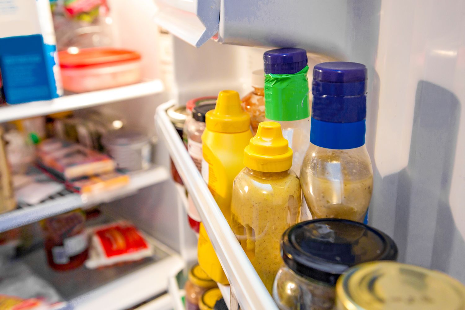 Condiments arranged on the inside door shelf of a refrigerator including mustard bottles and jars with other food items visible on shelves