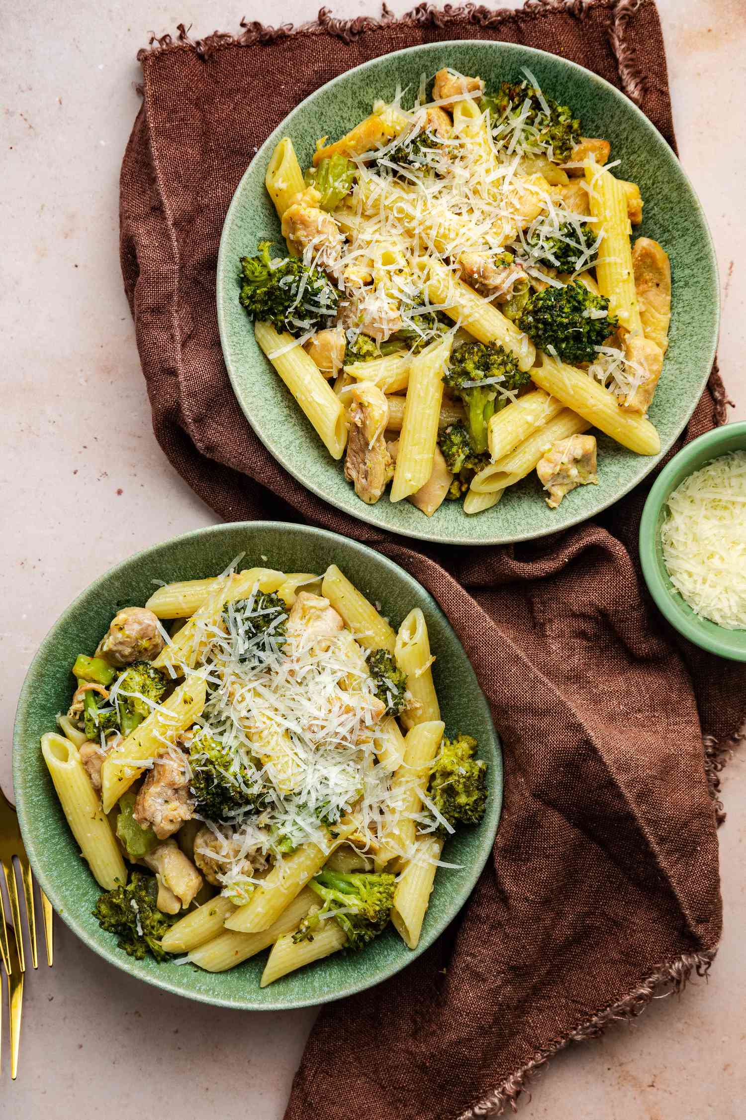 Two Bowls of Chicken and Broccoli Pasta topped with Parmesan Cheese on a Kitchen Towel and Next to a Small Bowl of Cheese and Some Utensils