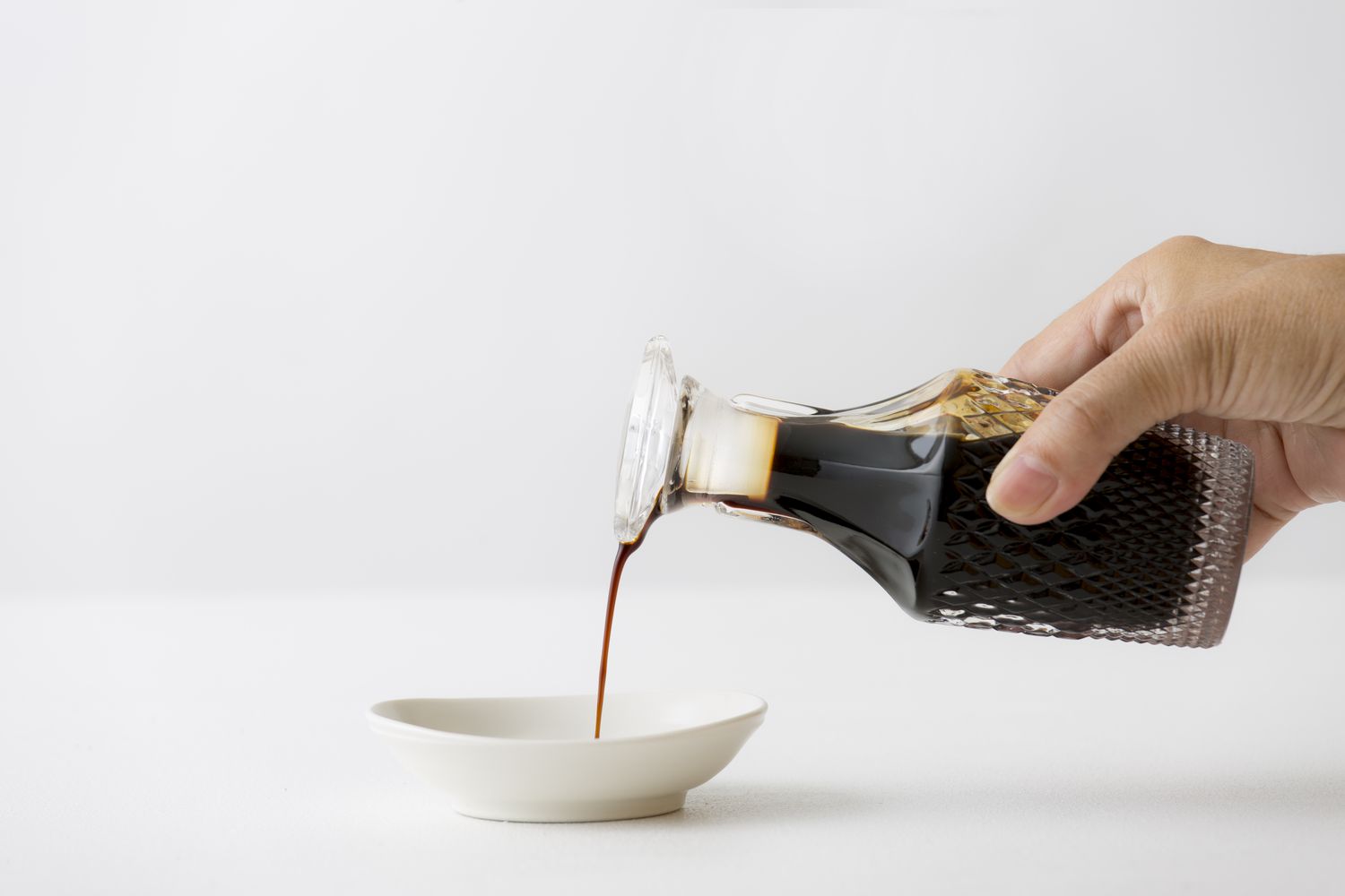 A hand holding a glass bottle pouring out soy sauce into a small sauce dish