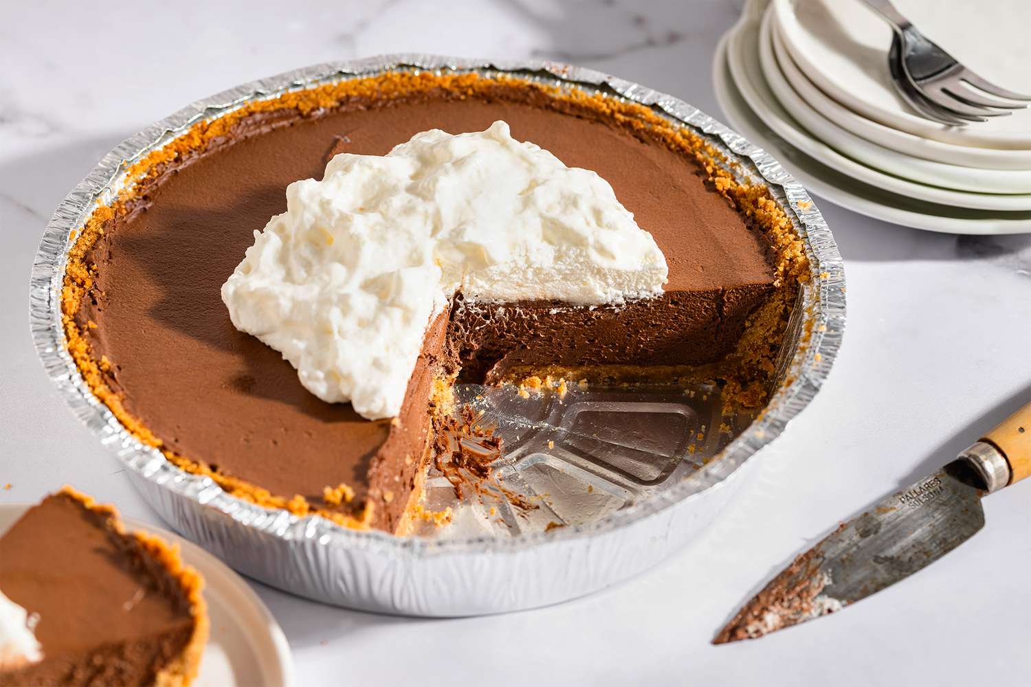 A chocolate cream pie with a slice removed on a table next to a knife and stack of plates