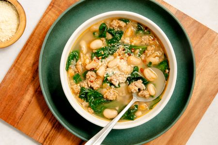 A white bowl with kale, white bean and sausage soup, places on a dark green plate on a wooden serving board