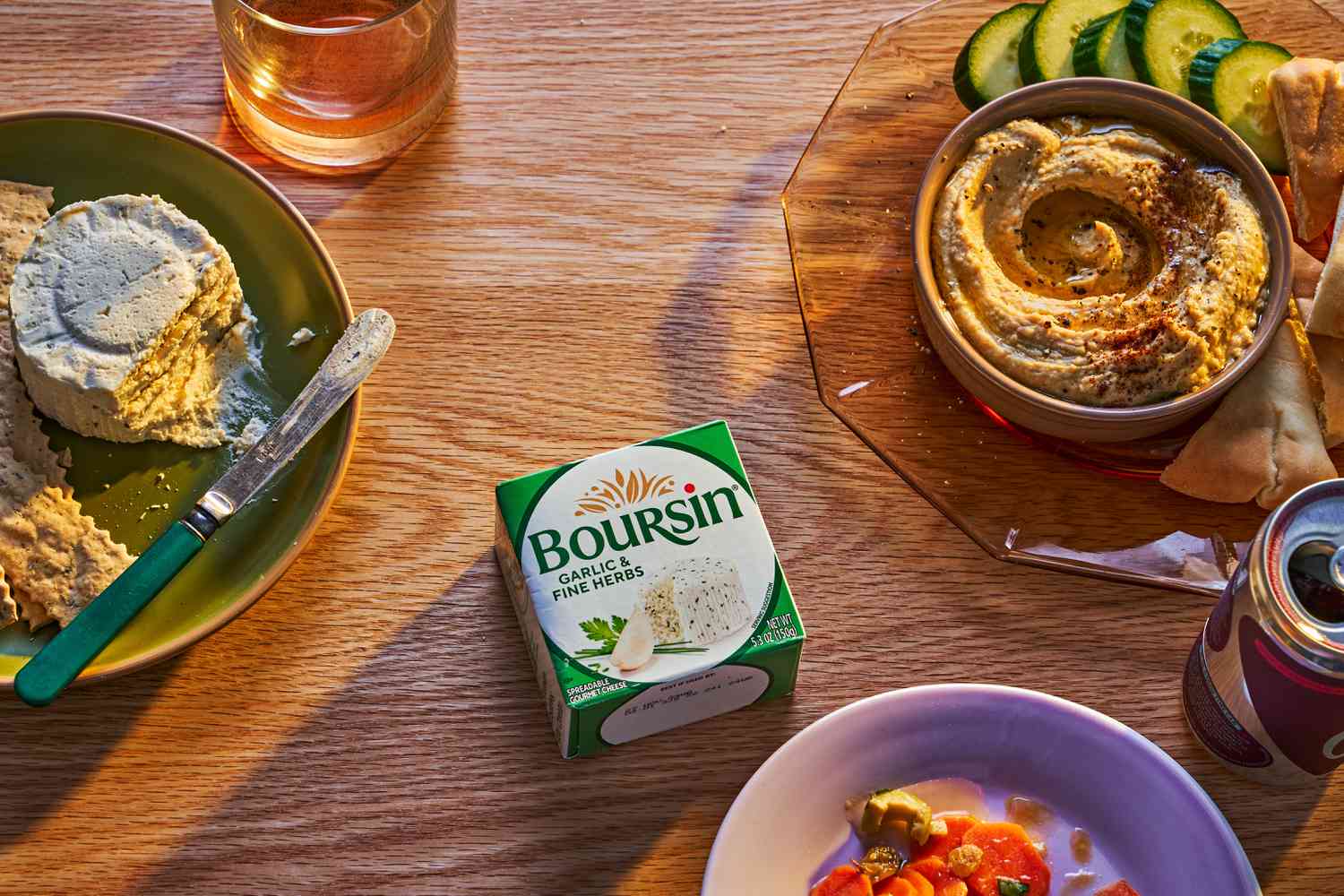 A coffee table surface with snacks like hummus, carrots, cucumbers, and Boursin cheese, including a branded box of Boursin