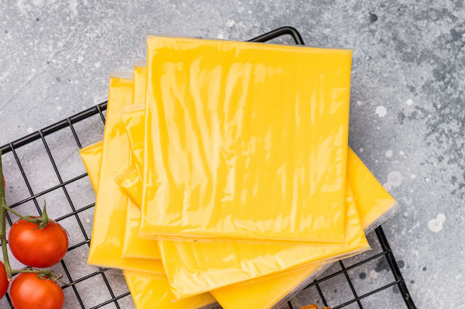 Slices of processed cheese in plastic wrapping on a grill rack with cherry tomatoes nearby