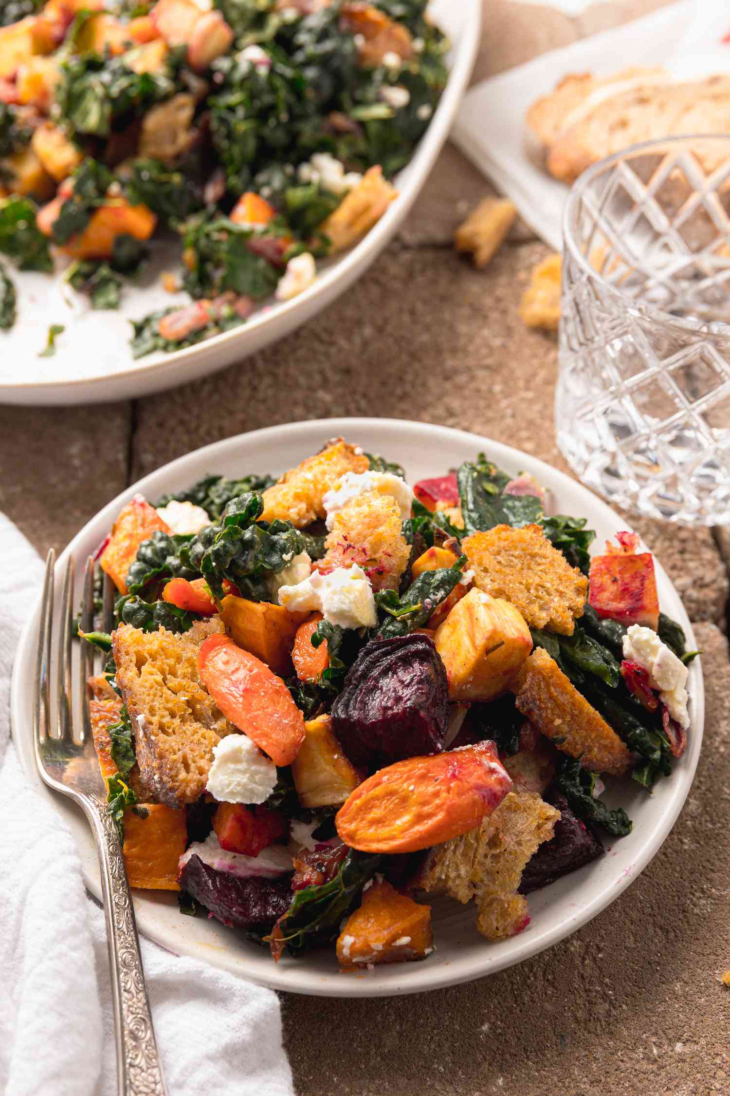 Serving of Roasted Root Vegetable Panzanella in a Bowl with a Fork, Surrounded by a Table Setting with a Platter of More Panzanella, a Glass of Water, and a Table Napkin,