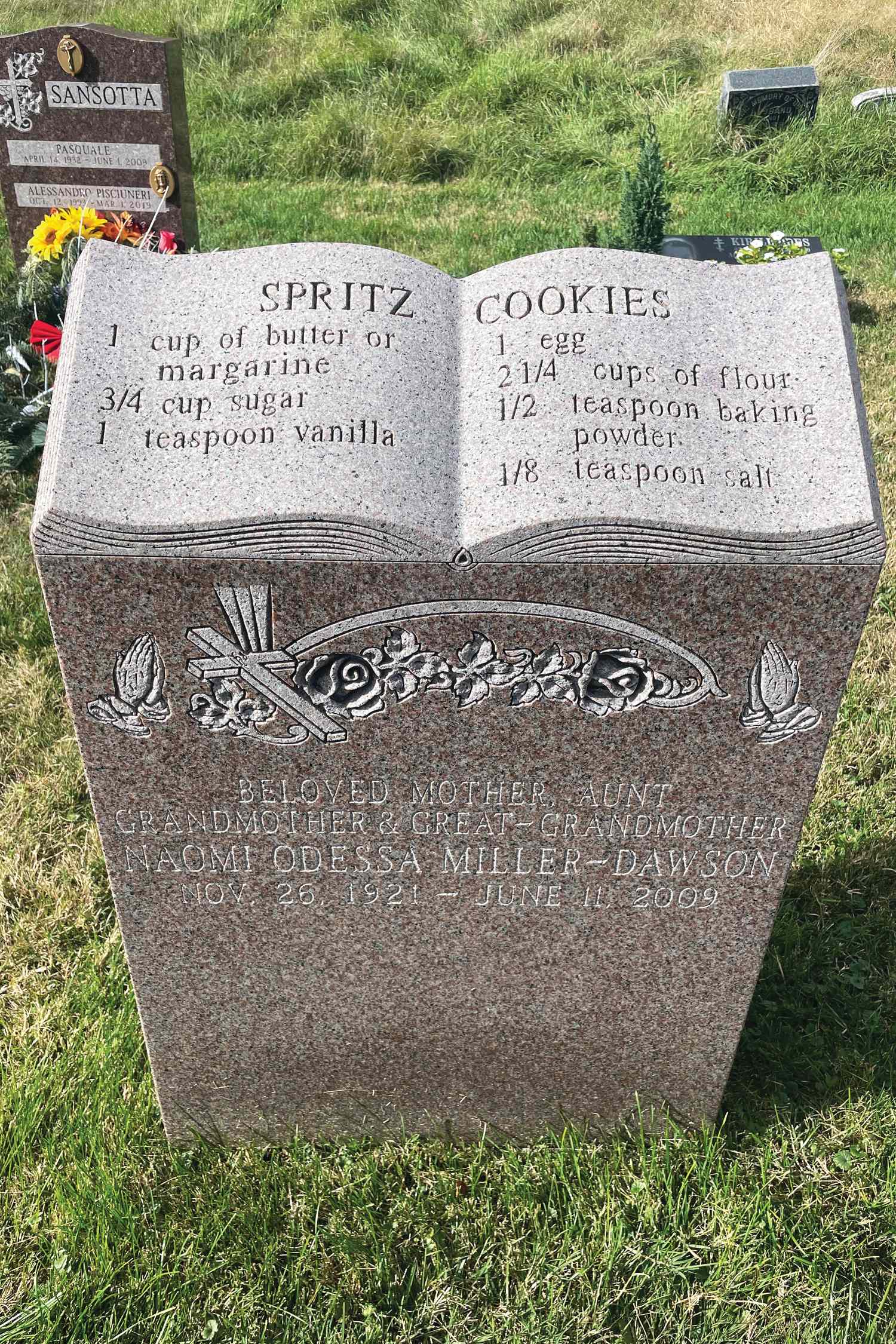 Tombstone engraved with a spritz cookie recipe resembling an open book surrounded by flowers in a cemetery