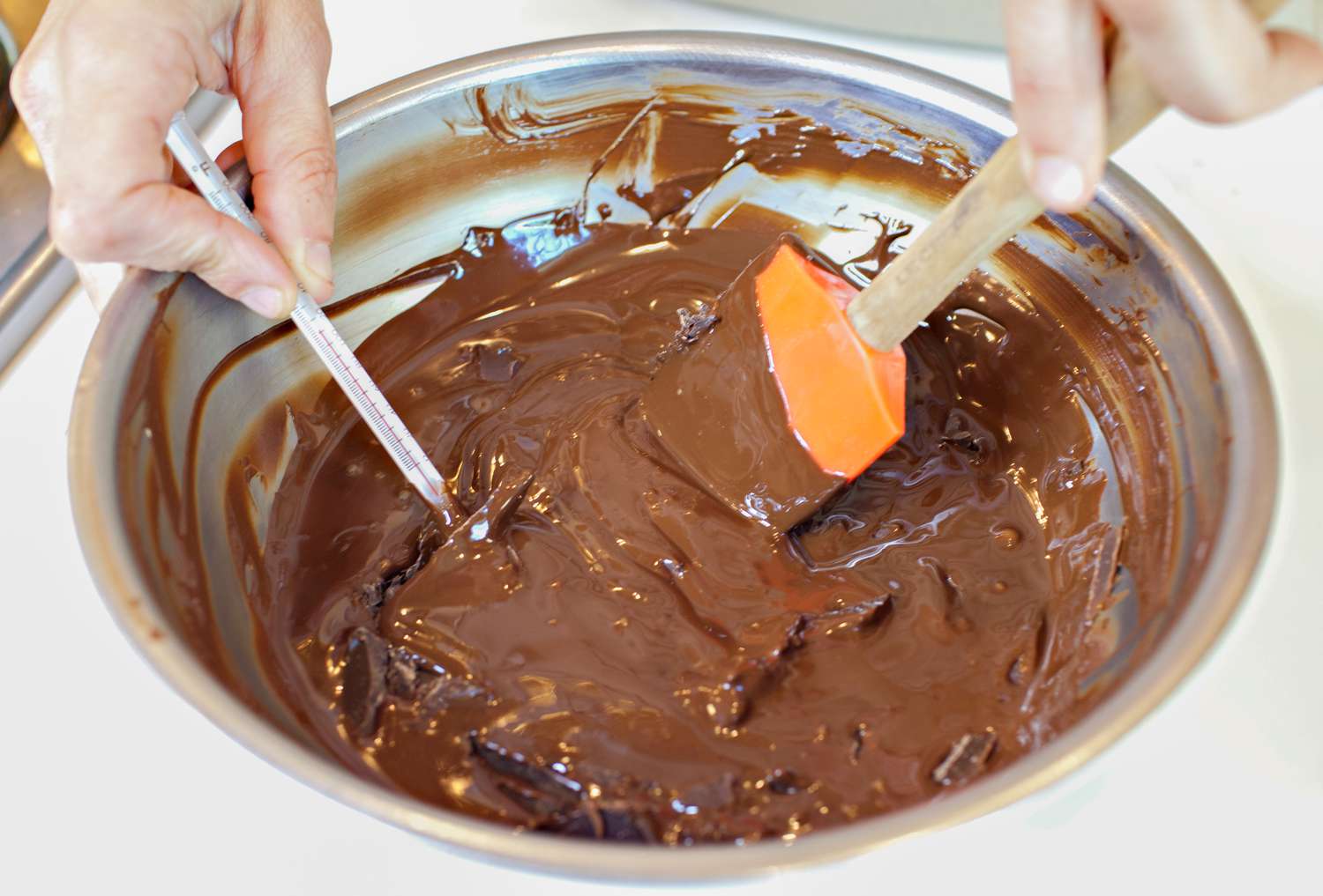 One Hand Holding Thermometer in Bowl of Melted Chocolate While Other Hand Stirs Chocolate Using a Rubber Spatula for How to Temper Chocolate