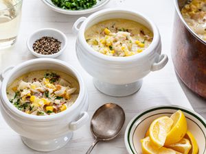 Corn and Crab Chowder in Two Soup Bowls with Handles at a Table Setting with a Pot with More Chowder, a Glass of White Wine, a Plate of Parsley, Small Bowl of Cracked Pepper, and Bowl of Lemon Slices