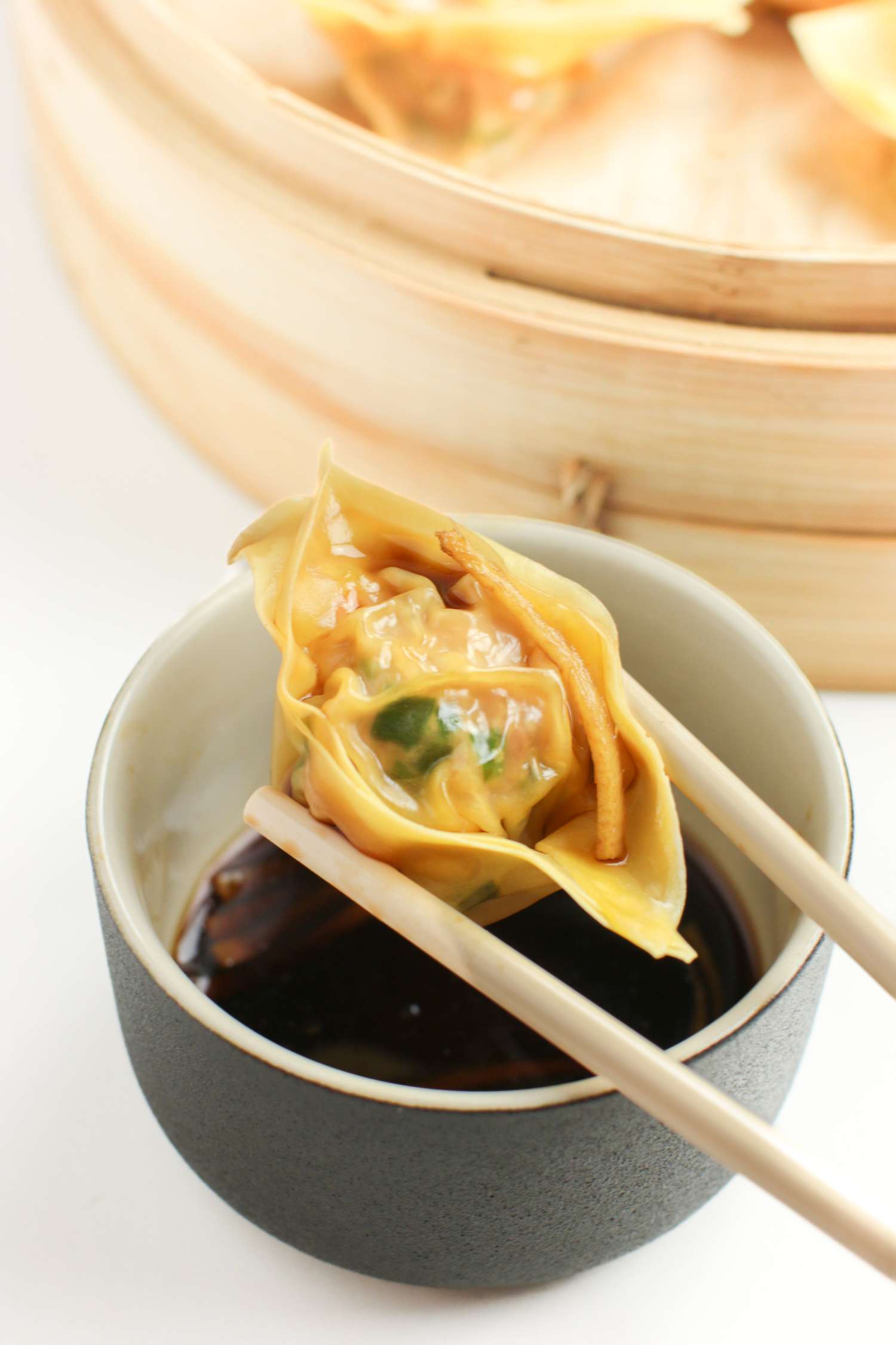 Pork and Chive Dumpling Held by a Pair of Chopsticks over a Small Bowl of Chinese Vinegar and Next to a Steamer